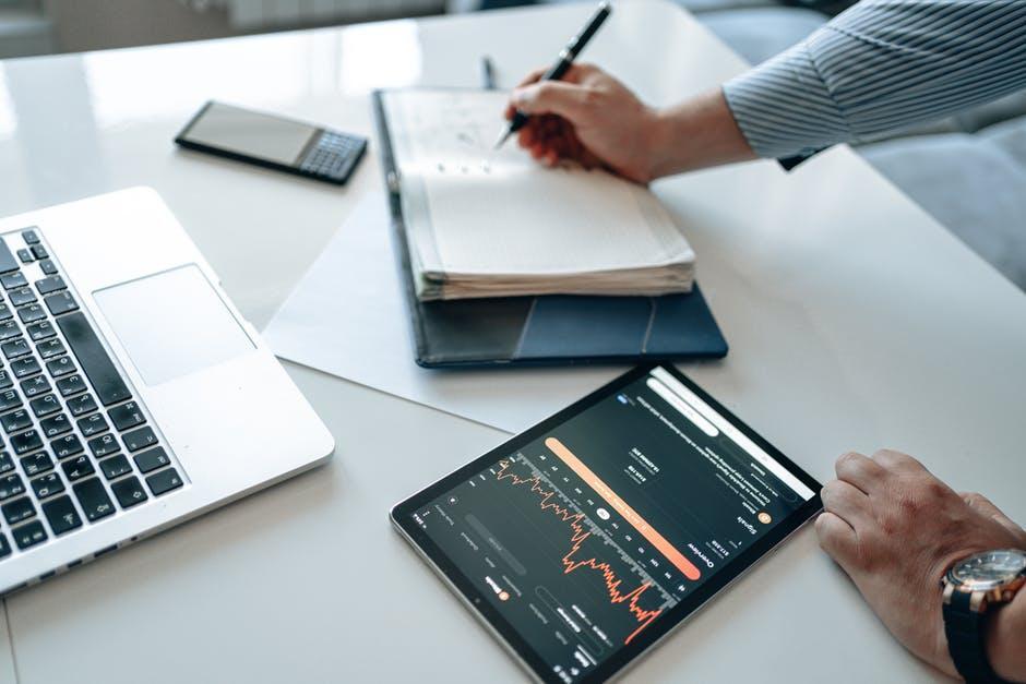 Person reviews stock market data on a tablet while taking notes at a clean white desk with a laptop and notebook nearby