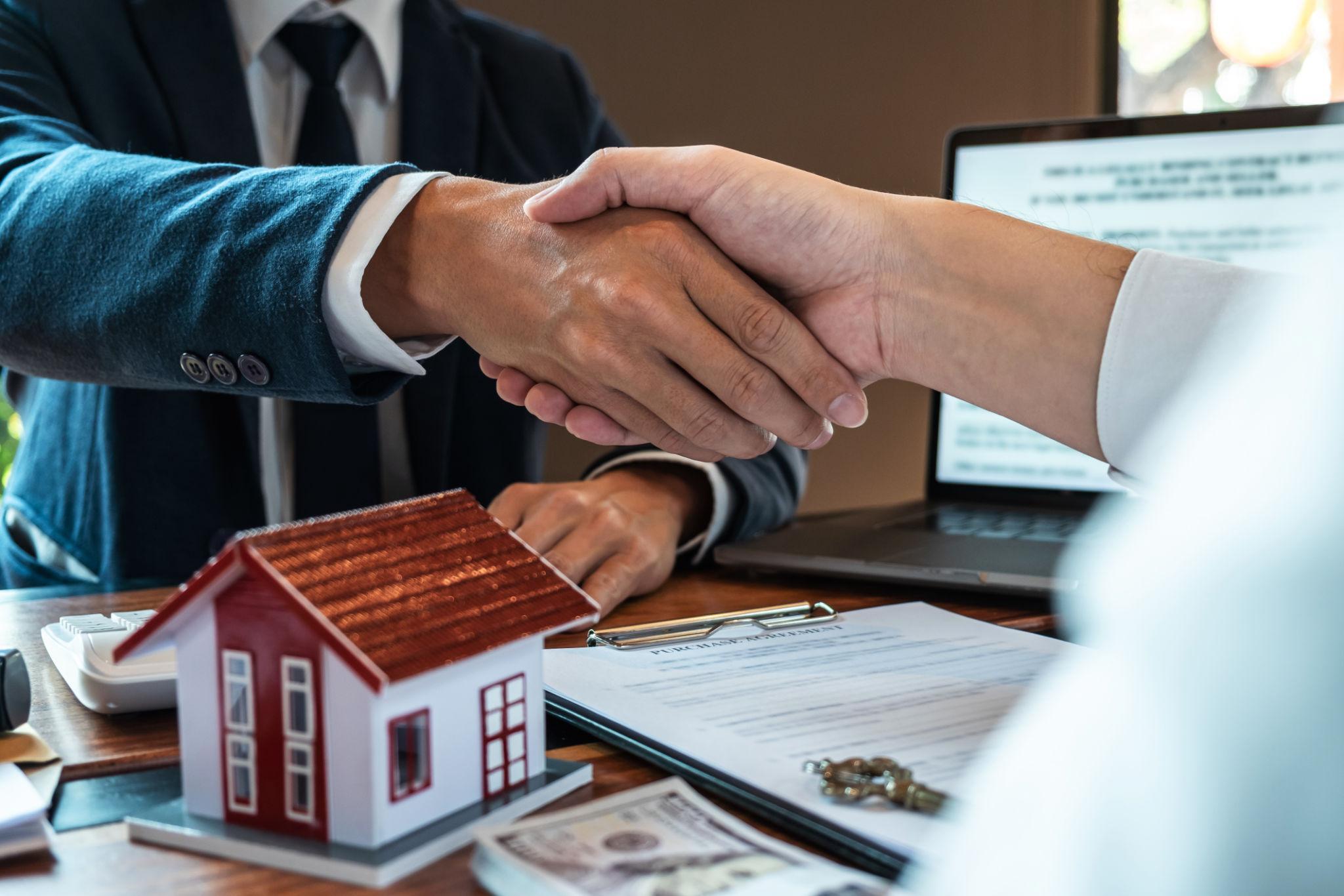 Two professionals shake hands across a desk, with a laptop, papers, and small house models nearby, signaling a real estate deal or agreement.