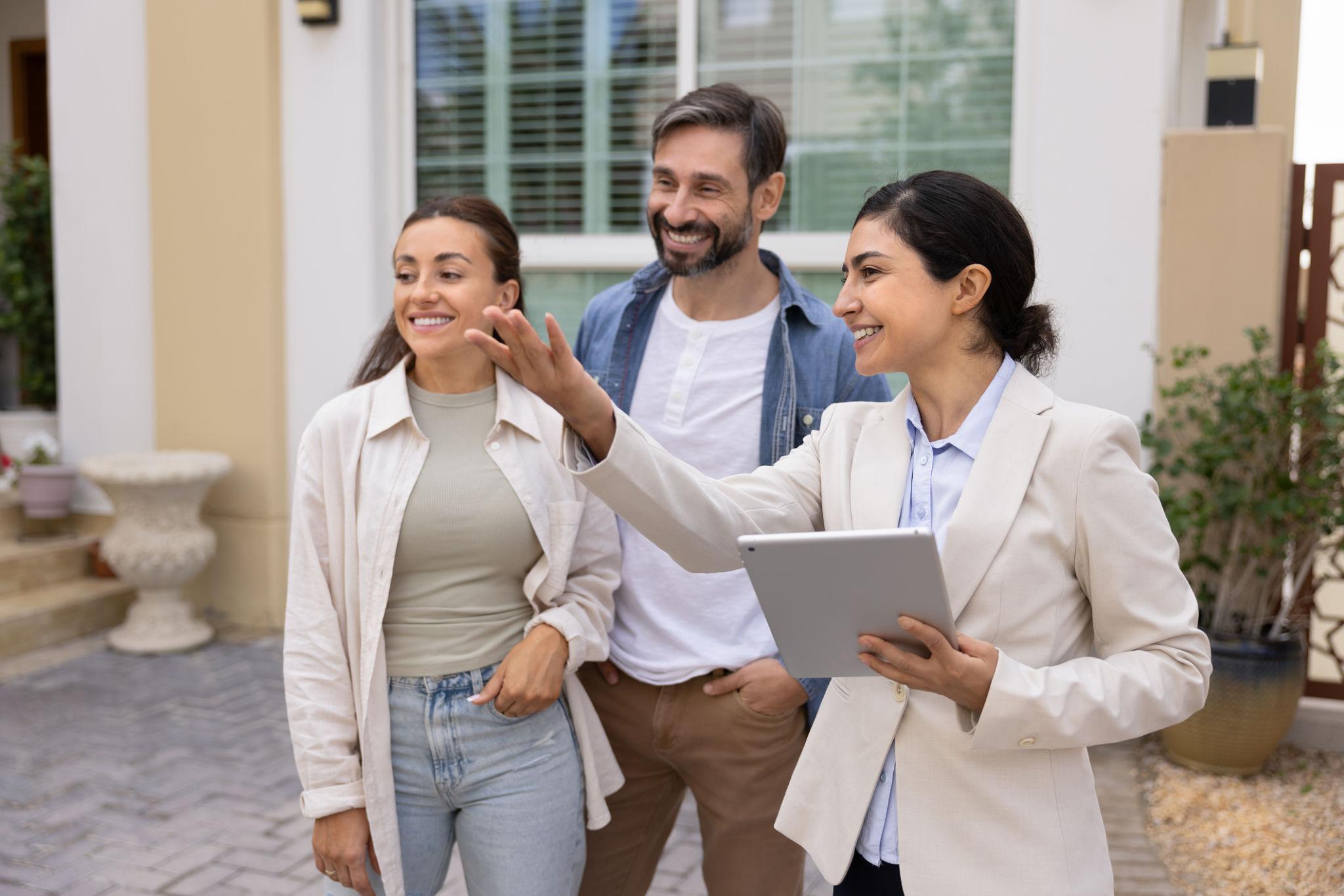 Three adults smiling outdoors in front of a modern house; a woman in a beige blazer holds a tablet and waves as two friends stand beside her.